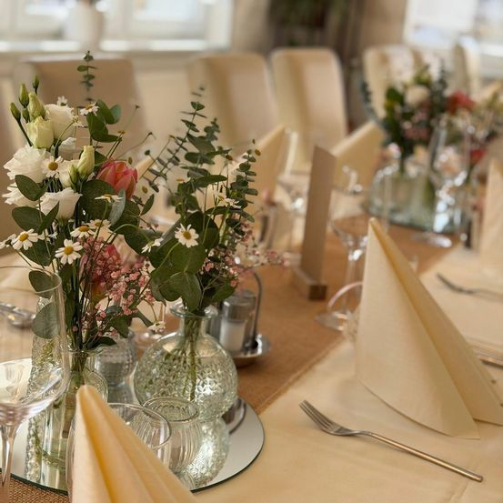Ein Tisch ist mit Blumen in Vasen, Silberbesteck, Gläsern und gefalteten Servietten gedeckt. Der Tisch ist mit einem beigen Tuch bedeckt.