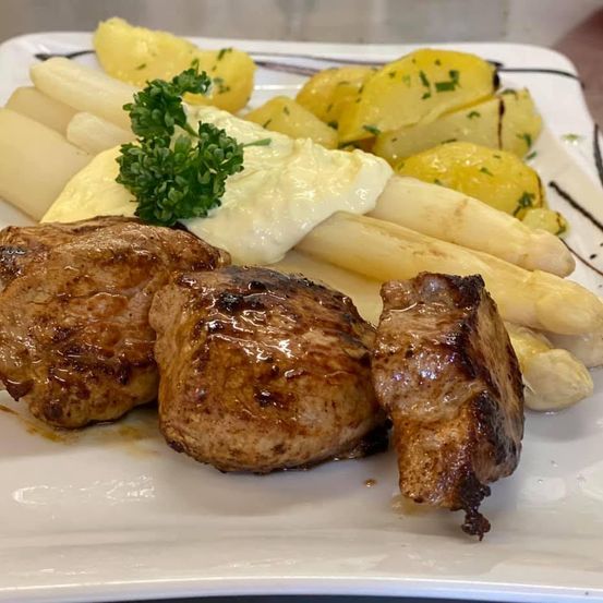 Ein Teller mit drei gegrillten Fleischstücken, weißem Spargel, Kartoffeln und einem Klecks Sahne. Das Fleisch ist mit Petersilie garniert.