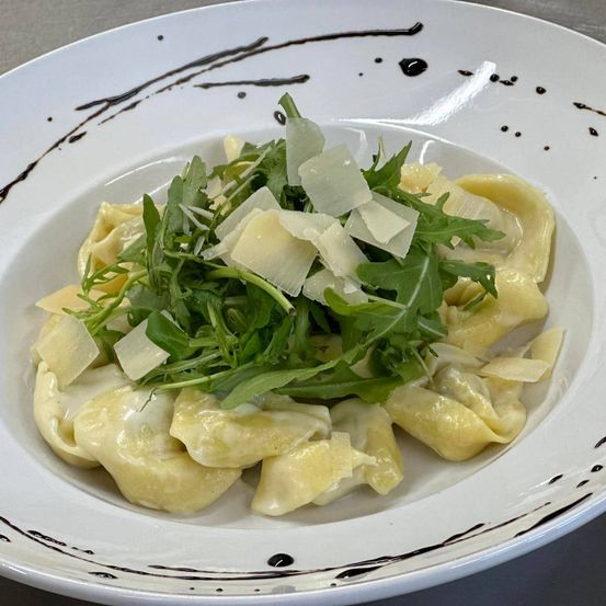 Ein weißer Teller mit Pasta, Rucola und geriebenem Käse darauf. Der Teller hat schwarze Linien und Punkte.