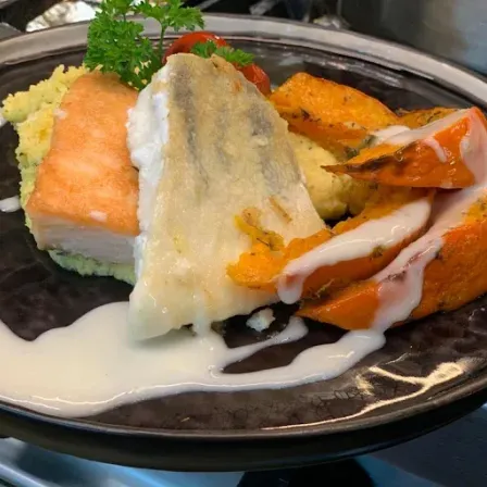 Ein Teller mit einem Stück Fisch, gelbem Reis und orangefarbenen Karotten, garniert mit einem grünen Kraut und mit weißer Sauce übergossen.