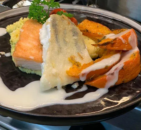 Ein Teller mit einem Stück Fisch, gelbem Reis und orangefarbenen Karotten, garniert mit einem grünen Kraut und mit weißer Sauce übergossen.