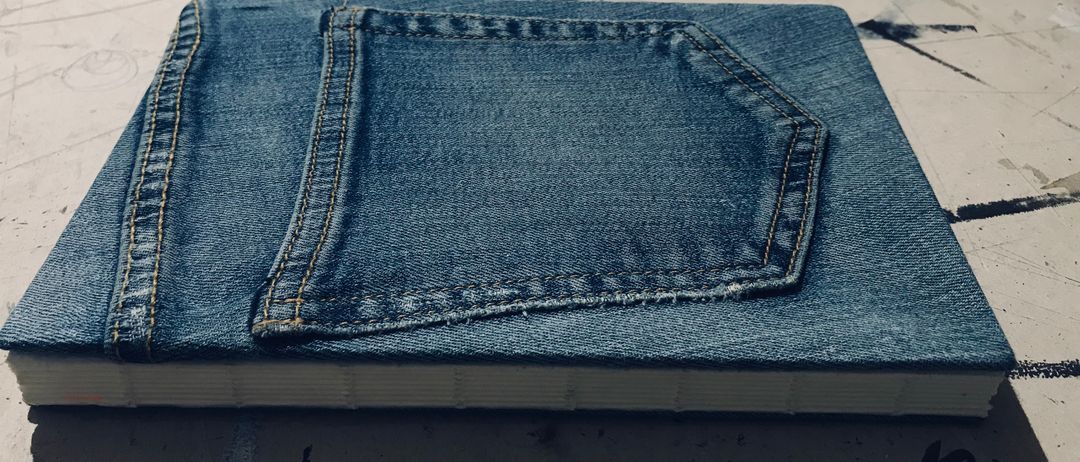 Ein Buchdeckel aus Denim mit sichtbarer Tasche auf der Vorderseite. Ein weißer Beutel mit dem Wort 'MEISTERBUCH' befindet sich dahinter. Der Denim ist blau und ordentlich gefaltet.