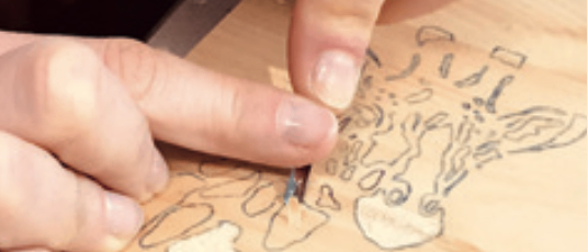 Eine Person zeichnet ein Zebra auf einem Holzbrett mit einem Werkzeug. Die Hände der Person sind sichtbar und die Zeichnung ist unvollständig.