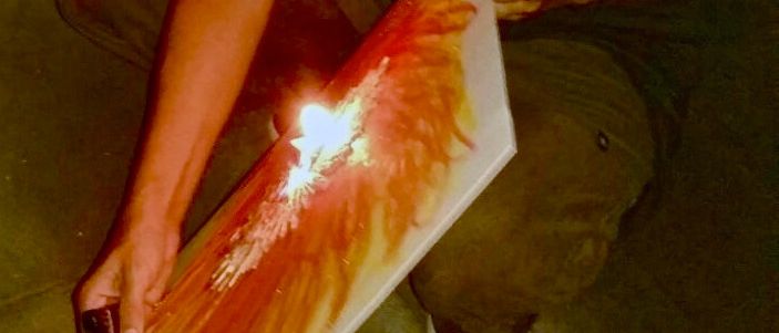 Eine Frau kniet auf dem Boden und hält ein Stück Papier mit einem brennenden Design. Sie ist in einem grünen ärmellosen Oberteil und Hose gekleidet, und ihre Haare sind zerzaust. Sie scheint sich in einem schwach beleuchteten Raum zu befinden.