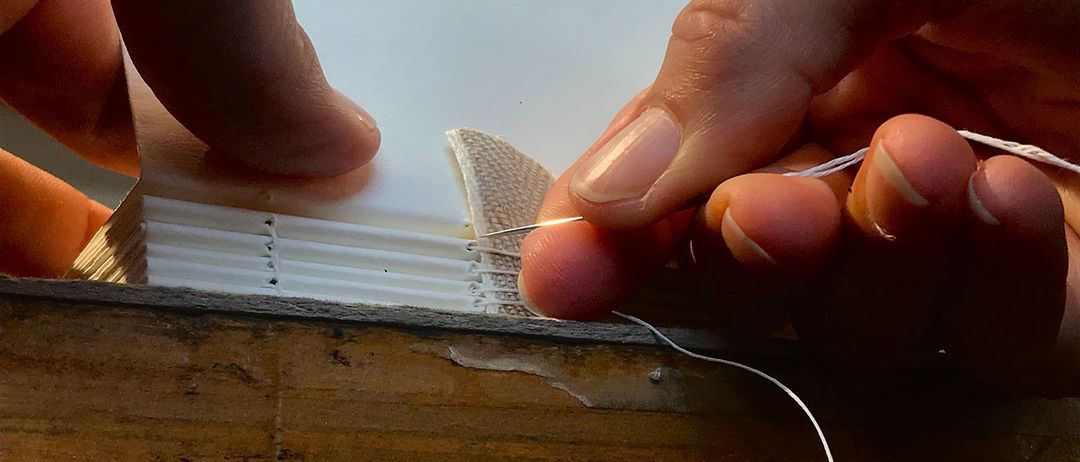 Eine Hand, die eine Nadel hält, fädelt einen weißen Stoffstreifen auf einem Holztisch ein. Der Stoff hat kleine Löcher, und die Nadel ist nah am Stoff.