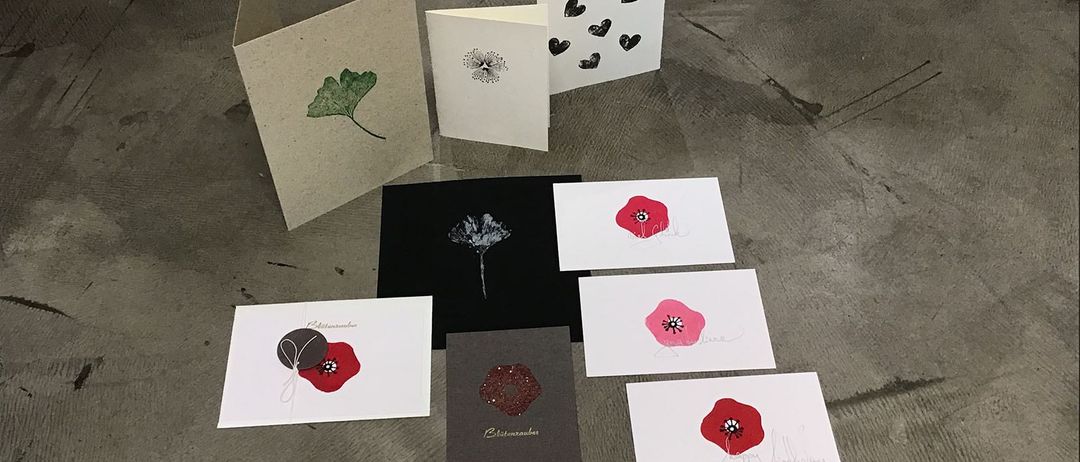 Sieben Grußkarten liegen auf einem Betonboden. Jede Karte hat ein anderes Design, darunter ein Ginkgoblatt, Herzen und Mohnblumen.