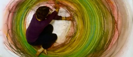 Eine Frau kniet inmitten einer großen, farbenfrohen Spirale auf dem Boden. Sie trägt ein lilafarbenes Shirt und schwarze Hose. Die Spirale besteht aus leuchtenden Farben wie Grün, Gelb und Rot.