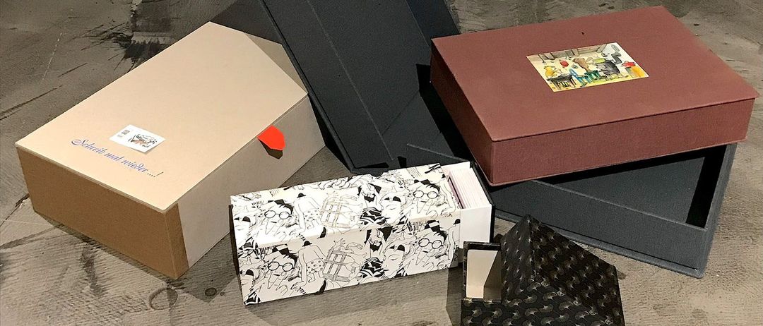 Eine Sammlung verschiedener Boxen wird auf einem Boden ausgestellt. Eine ist geöffnet, während die anderen geschlossen sind. Die offene ist schwarz und hat ein Grafik darauf.