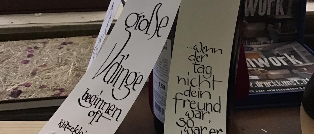 Zwei gefaltete weiße Papiere mit schwarzer Handschrift werden auf einem Tisch präsentiert. Das linke Papier zeigt 'Vinge beginn offen' und das rechte 'Wenn der tag nicht dein freund war, so war er dein lehrer'.