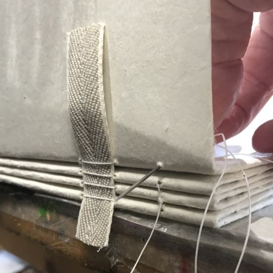 Ein Nahaufnahme einer Hand, die ein Buch mit einer Nadel näht, gesichert durch ein graues Band. Das Buch besteht aus mehreren gefalteten weißen Seiten, die auf einem Holztisch liegen.