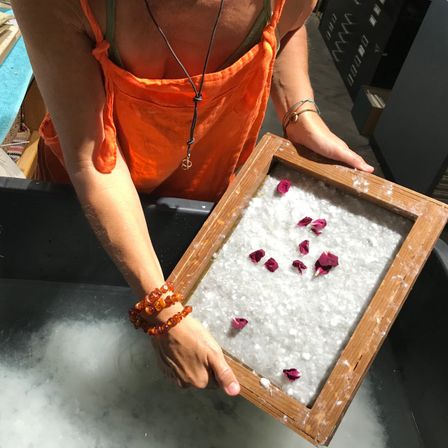 Eine Frau in einem orangefarbenen Kleid hält ein Holzrahmen mit Rosenblättern in einer Badewanne.