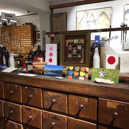 Ein Raum gefüllt mit verschiedenen Kunstwerken, darunter Gemälde, eine Vase und ein Holzschrank. Auf dem Tisch liegen Postkarten, eine herzförmige Karte und ein gerahmtes Bild. An der Wand hängt ein großes Gemälde. Der Raum hat Holzschränke und Regale.