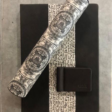 Eine aufgerollte Yogamatte mit aufwendigen Mustern liegt auf einer schwarz-weiß gestreiften Oberfläche. Daneben befindet sich ein kleiner schwarzer Kasten mit dem Wort 'Godi'. Der Hintergrund ist ein strukturierter Betonboden.