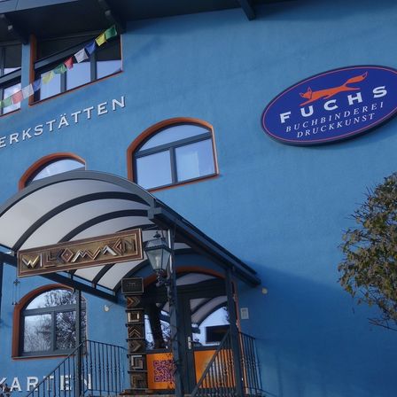 Ein blaues Gebäude mit Rundbogenfenstern und einem Schild, das 'Universal Grafische Werkstatten' besagt. Ein überdachter Eingang mit dem Schild 'Wiloma' führt zum Gebäude. Flaggen hängen über den Fenstern.