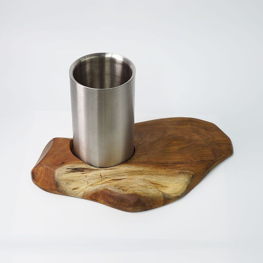 Ein silberner Edelstahlbecher steht auf einer hölzernen Unterlage vor weißem Hintergrund. Die Unterlage ist unregelmäßig geformt und aus Holz.