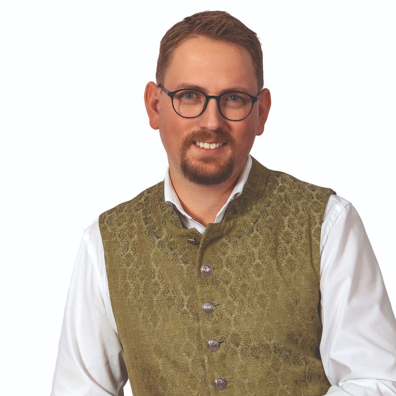Ein Mann mit Bart und Schnurrbart in einem weißen Hemd und einem gemusterten grünen Westchen posiert für ein Foto.