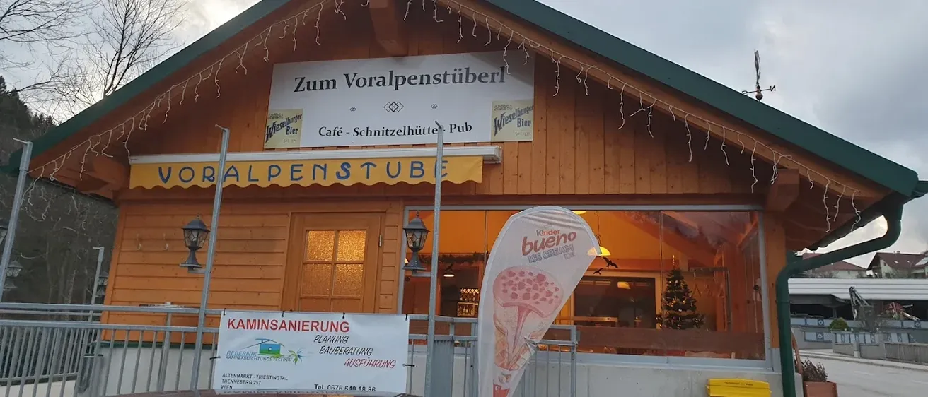 Ein Gebäude mit einem Schild, das Zum Voralpenstuberl Cafe - Schnitzelhütte Pub anzeigt. Unten am Schild befindet sich ein Banner mit Kaminsanierung Planung Bauberatung Ausführung. Es gibt auch ein Schild mit Kinder Bueno Eiscreme. Rechts nebenan steht ein Weihnachtsbaum.