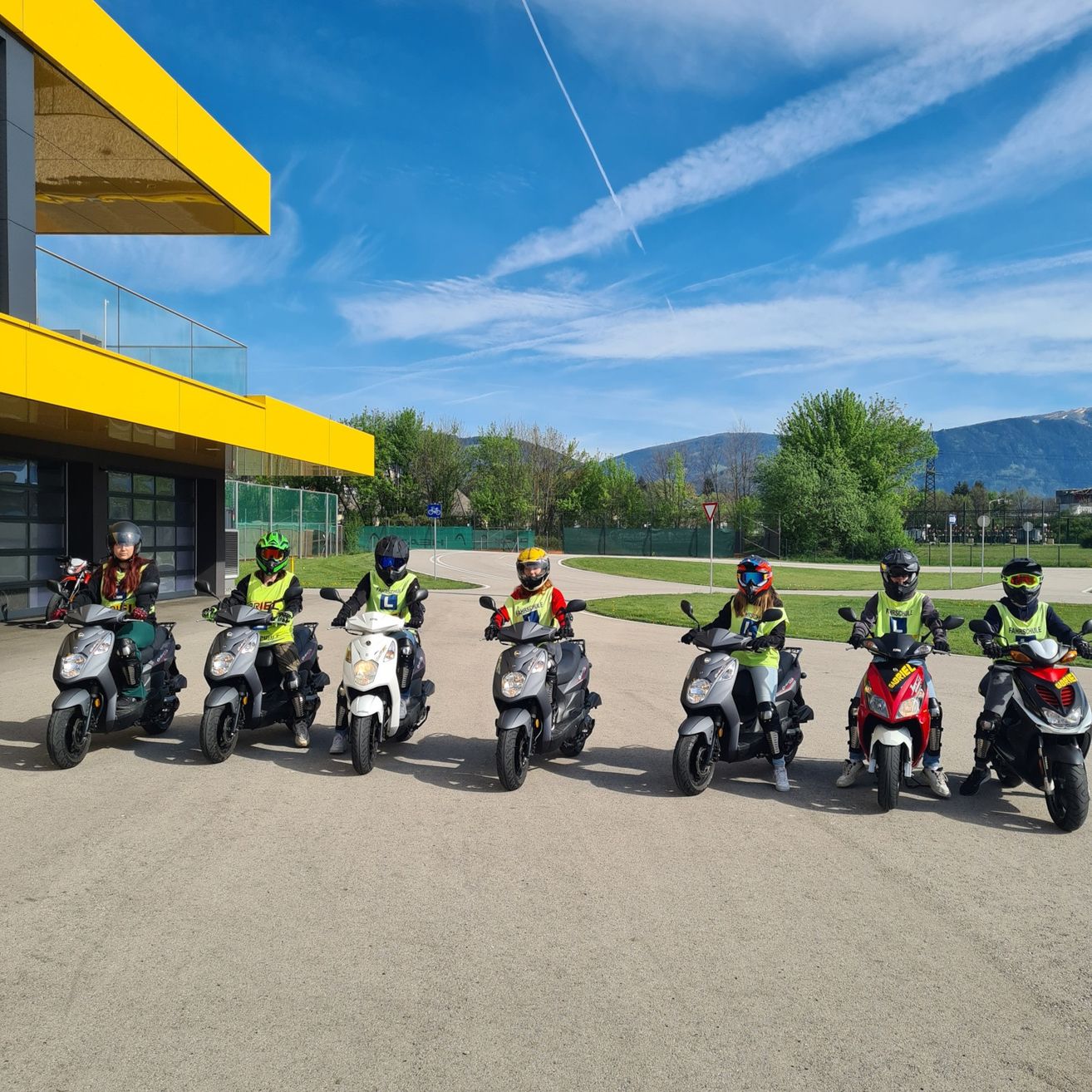 Eine Gruppe von Menschen in Schutzkleidung steht mit ihren Rollern vor einem gelben Gebäude unter einem blauen Himmel mit Wolken und Bergen in der Ferne.