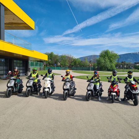 Eine Gruppe von Menschen in Schutzkleidung steht mit ihren Rollern vor einem gelben Gebäude unter einem blauen Himmel mit Wolken und Bergen in der Ferne.