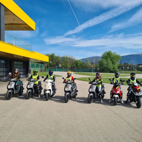 Eine Gruppe von Menschen in Schutzkleidung steht mit ihren Rollern vor einem gelben Gebäude unter einem blauen Himmel mit Wolken und Bergen in der Ferne.