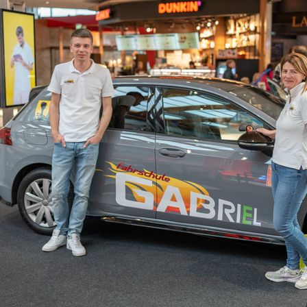 Ein Mann und eine Frau stehen neben einem Auto in einem Einkaufszentrum. Das Auto hat das Wort Gabriel an der Seite. Dahinter befindet sich ein Geschäft mit einem Schild, das Dunkin lautet.