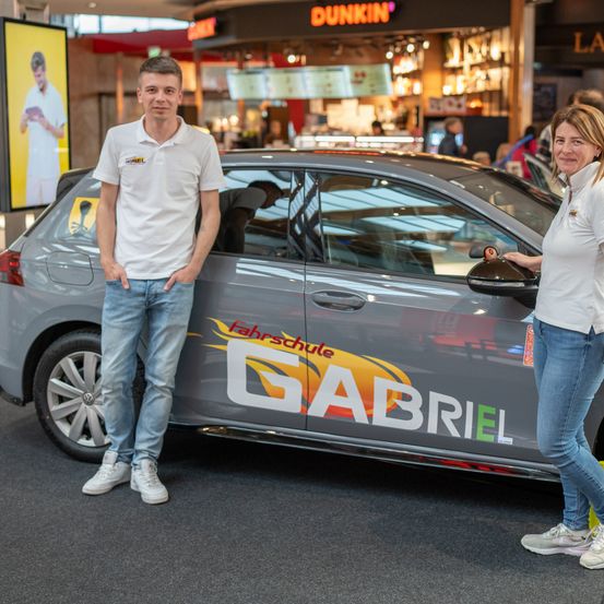 Ein Mann und eine Frau stehen neben einem Auto in einem Einkaufszentrum. Das Auto hat das Wort Gabriel an der Seite. Dahinter befindet sich ein Geschäft mit einem Schild, das Dunkin lautet.