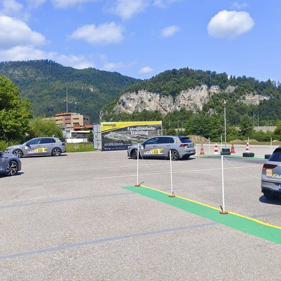 Ein Parkplatz mit mehreren geparkten Autos, einer Bergkette im Hintergrund und einem Banner mit dem Text 'Fahrsicherheits-training'. Es gibt Verkehrskegel und Barrieren auf dem Boden.
