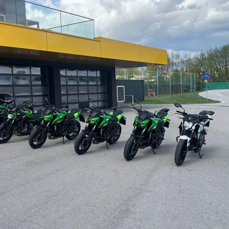 Mehrere grüne und schwarze Motorräder in einer Reihe vor einem Gebäude mit einem gelben Vordach geparkt. Bäume und ein Zaun sind im Hintergrund zu sehen.