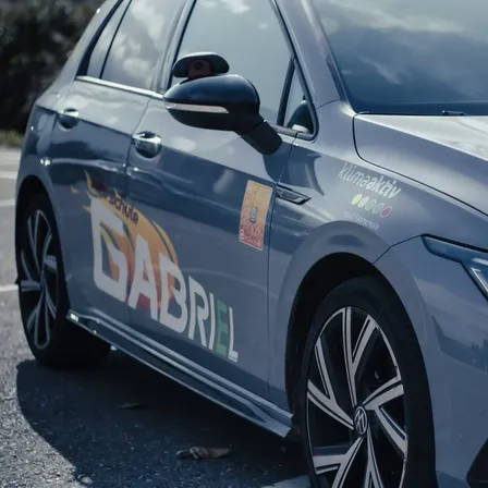 Ein silberner Wagen mit einem Aufkleber mit dem Namen 'Gabriel' und verschiedenen Logos steht am Straßenrand. Der vordere Stoßfänger des Autos hat eine Lampe.