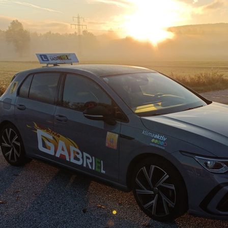 Ein graues Auto mit dem Wort 'GABRIEL' an der Seite ist auf einem Kiesweg bei Sonnenuntergang geparkt, mit Nebel im Hintergrund und einem Feld mit Bäumen und Bergen im Blick.