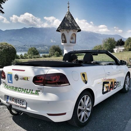 Ein weißer Cabriolet mit dem Wort Fahrschule an der Seite ist vor einer Kapelle geparkt.
