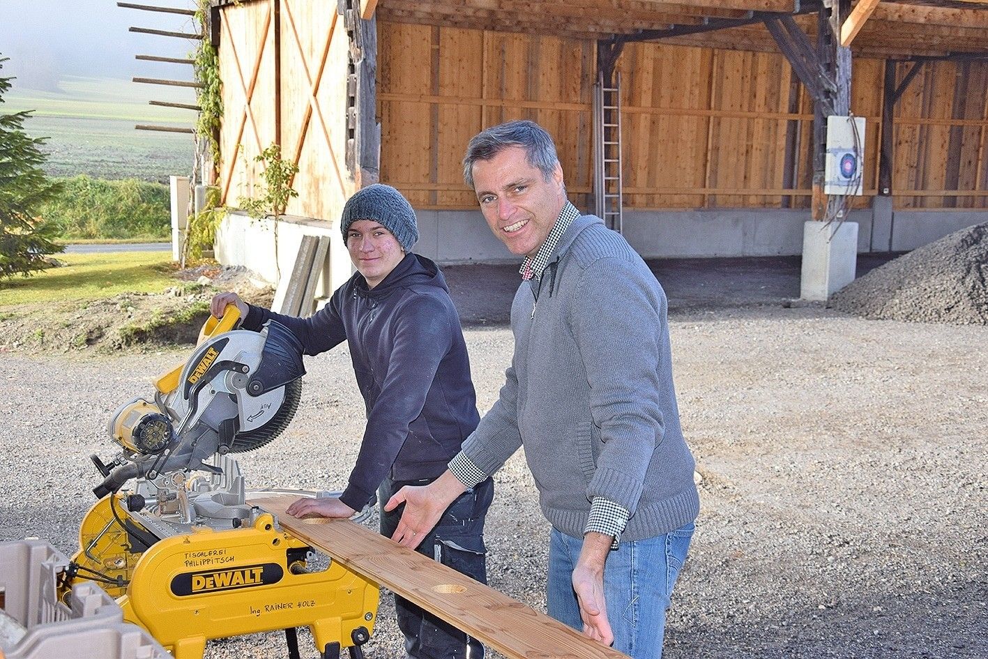 Zwei Männer arbeiten an einer Kreissäge, lächeln und posieren für ein Foto vor einer Holzscheune.