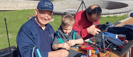 Drei Männer arbeiten draußen an einem Projekt auf einem Tisch. Ein Mann trägt ein blaues Shirt und einen Hut, während ein anderer Mann ein rotes Shirt trägt. Der dritte Mann benutzt ein Löteisen.