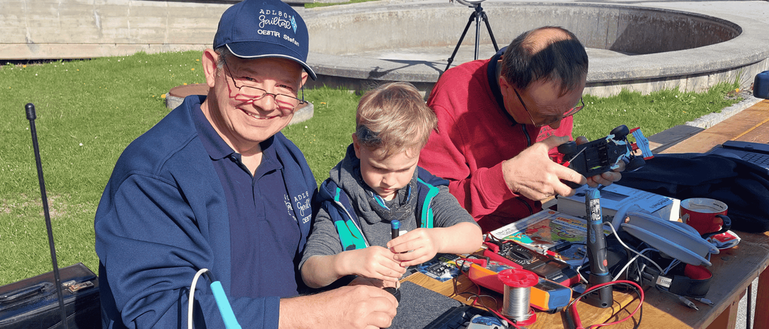 Drei Männer arbeiten draußen an einem Projekt auf einem Tisch. Ein Mann trägt ein blaues Shirt und einen Hut, während ein anderer Mann ein rotes Shirt trägt. Der dritte Mann benutzt ein Löteisen.