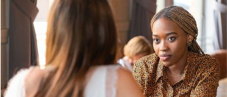 Eine Frau mit geflochtenen Haaren, die ein Leopardenmusterhemd trägt, schaut eine verschwommene Frau mit langen Haaren an. Dahinter ist ein verschwommenes Bild einer weiteren Frau zu sehen.