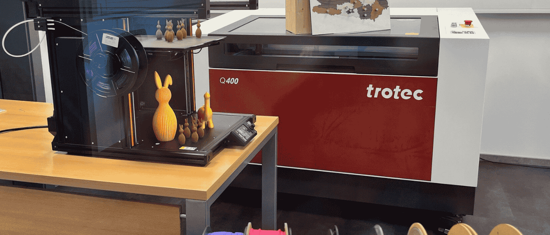Ein 3D-Drucker mit einem Holzkasten und einer Karte oben, umgeben von Spulen mit farbigem Filament. Der Drucker ist mit 'Q400' und 'trotec' beschriftet.