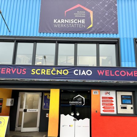 Das Gebäude hat eine blaue Außenseite mit Glasfenstern. Ein Banner über dem Eingang liest 'SERVUS SCREČNO CIAO WELCOME'. Der Eingang hat zwei Türen mit einem Schild, das 'Personal' steht. Es gibt einen Geldautomaten auf der rechten Seite des Eingangs.