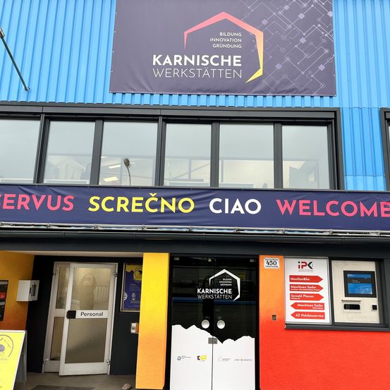 Das Gebäude hat eine blaue Außenseite mit Glasfenstern. Ein Banner über dem Eingang liest 'SERVUS SCREČNO CIAO WELCOME'. Der Eingang hat zwei Türen mit einem Schild, das 'Personal' steht. Es gibt einen Geldautomaten auf der rechten Seite des Eingangs.