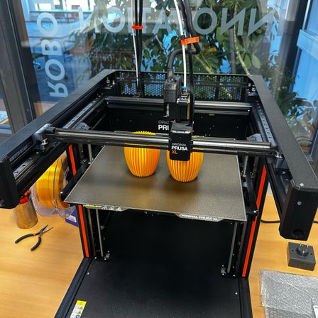 Ein Prusa XL 3D-Drucker ist zu sehen, mit zwei gelben gedruckten Objekten auf seiner Plattform. Der Drucker steht auf einem Holztisch mit sichtbarem Netzkabel.