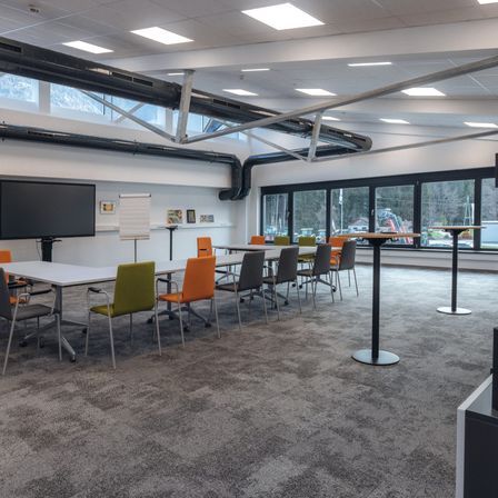 Ein leerer Besprechungsraum mit Tischen und Stühlen um einen zentralen Tisch herum angeordnet, mit einem großen Bildschirm und einer Whiteboard. Der Raum hat große Fenster und Metallbalken an der Decke.