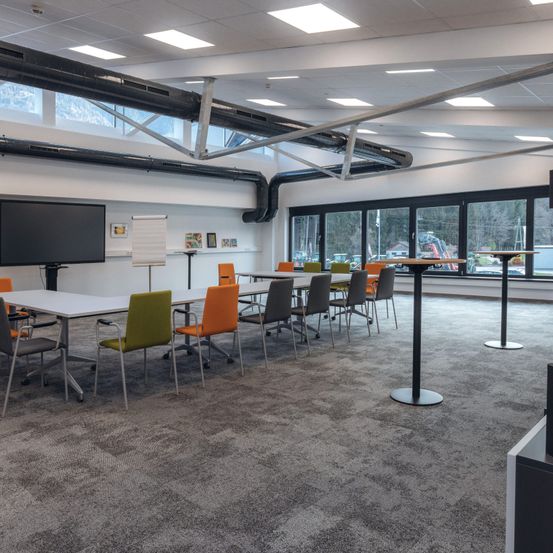 Ein leerer Besprechungsraum mit Tischen und Stühlen um einen zentralen Tisch herum angeordnet, mit einem großen Bildschirm und einer Whiteboard. Der Raum hat große Fenster und Metallbalken an der Decke.