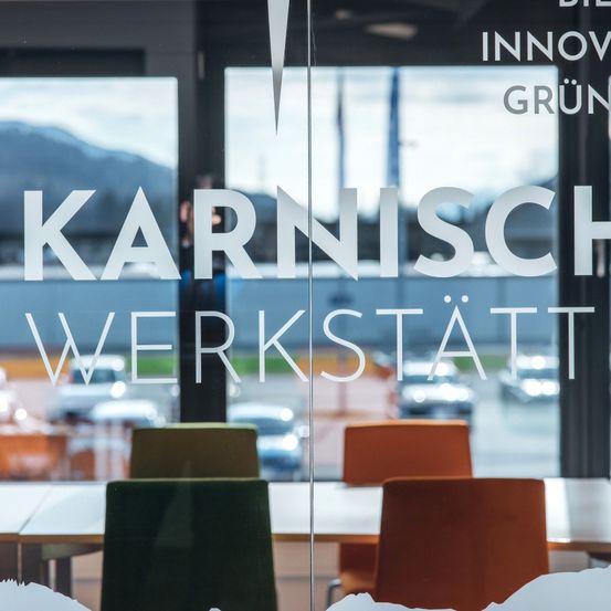 Eine Glaswand zeigt die Worte 'KARNISCH WERKSTATT' mit einer verschwommenen Ansicht eines Parkplatzes und Bergen im Hintergrund.