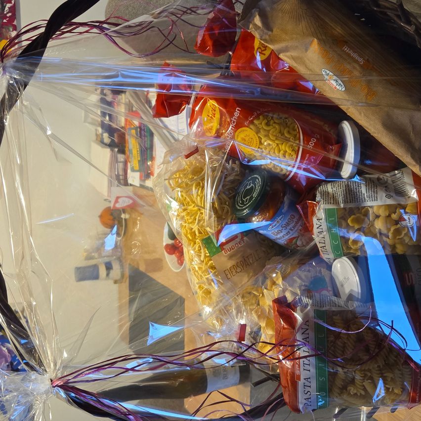 Ein durchsichtiger Plastikbeutel enthält verschiedene Lebensmittel wie Pasta, Dosentomaten und andere verpackte Waren. Der Beutel ist mit lila Bändern gebunden und auf einer Holzoberfläche platziert.