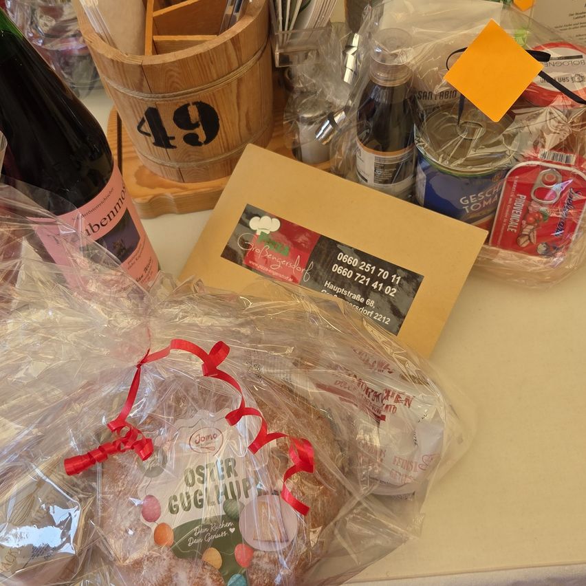 Auf einem Tisch sind ein verpacktes Geschenk, eine Flasche Wein und andere Gegenstände zu sehen. Das Geschenk ist in Plastik eingewickelt und hat ein rotes Band. Dahinter befindet sich ein hölzerner Eimer mit der Nummer 49. Andere Gegenstände sind eine Plastiktüte, eine Dose und ein gelber Haftnotizzettel.