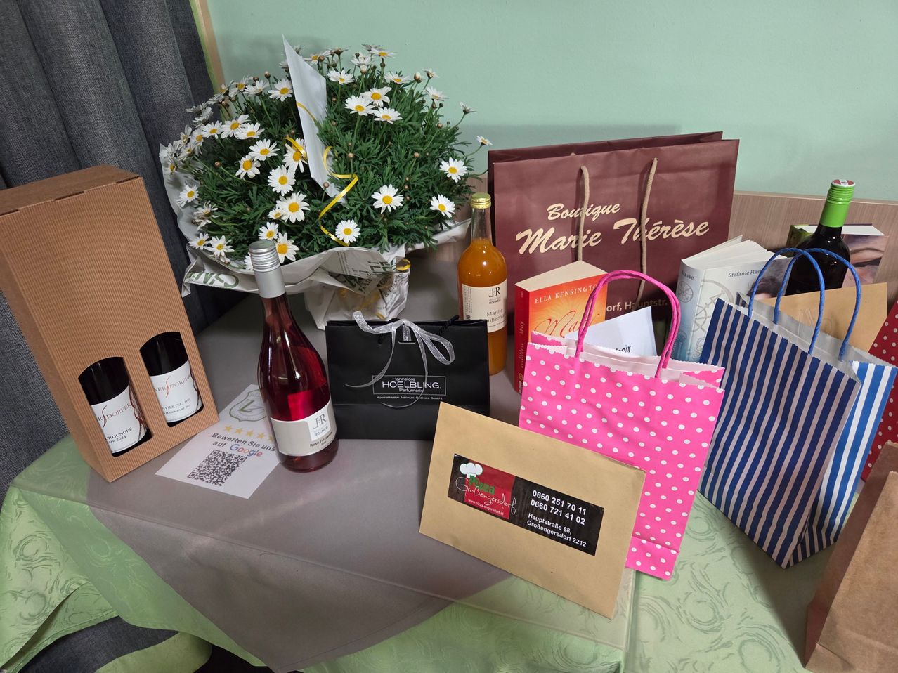 Ein Tisch zeigt einen Strauß Gänseblümchen, eine Flasche Wein, eine Parfümdose und verschiedene Einkaufstüten. Die Einkaufstüten umfassen eine rosa gepunktete Tasche und eine blau-weiß gestreifte Tasche. Die Parfümflasche hat ein Etikett.