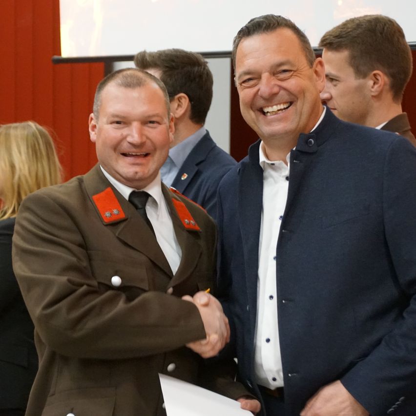 Zwei Männer in formeller Kleidung schütteln sich die Hände und lächeln vor einem verschwommenen Hintergrund.