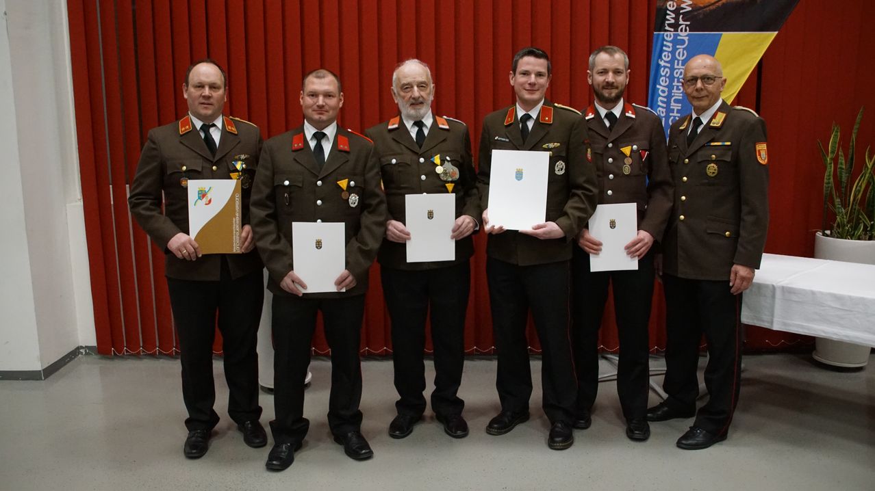 Fünf Männer in Militäruniformen stehen vor einem roten Vorhang und halten Zertifikate mit Logos und Text in den Händen. Sie lächeln und schauen in die Kamera.