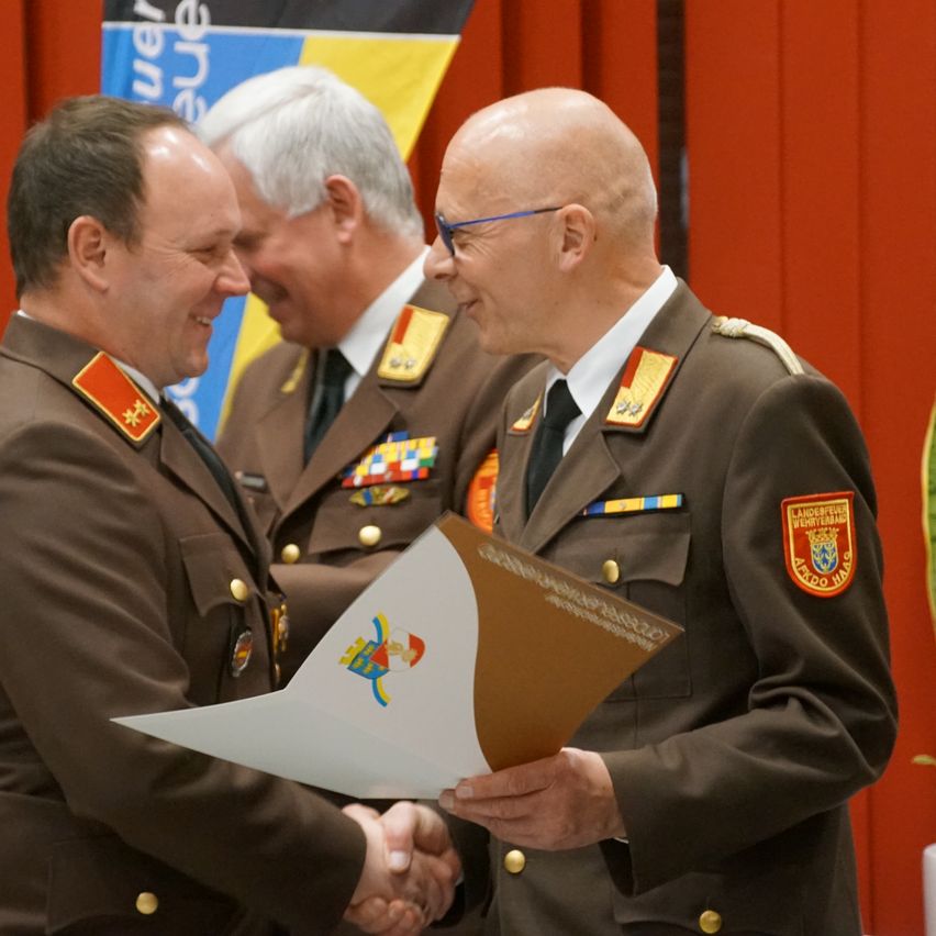 Drei Militäroffiziere in Uniform schütteln sich die Hände. Der Mann auf der rechten Seite trägt eine Brille. Hinter ihnen befindet sich ein roter Vorhang mit einer Flagge und einem gelben Rand.