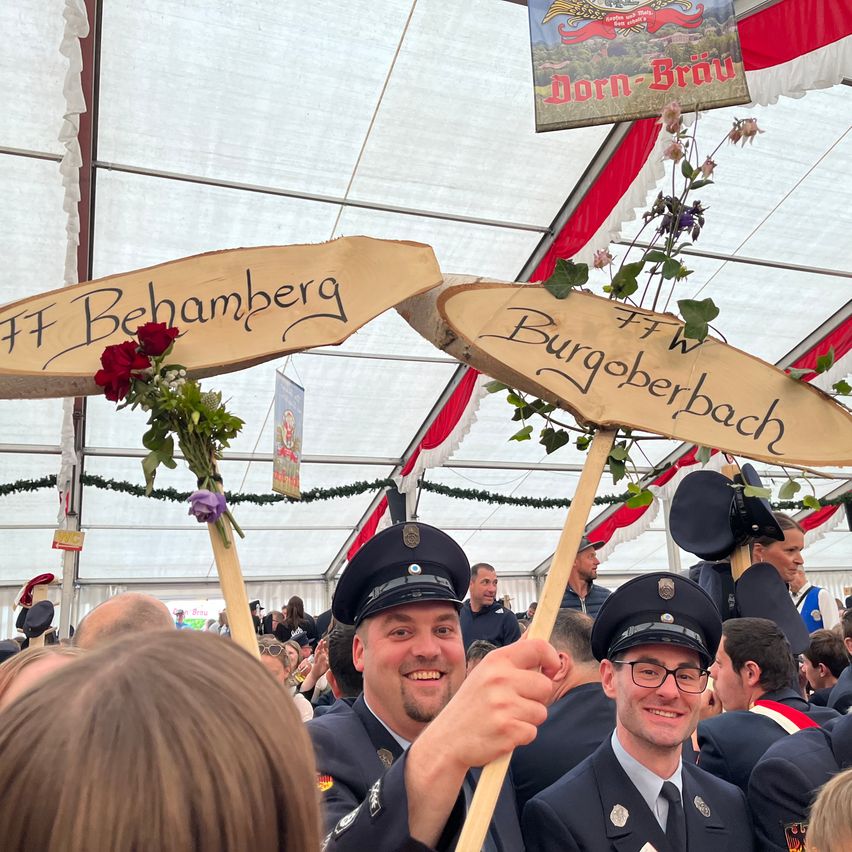 Zwei Männer in Uniform halten Schilder mit den Aufschriften FF Behamberg und FFW Burgoberbach, stehend in einer festlichen Menge, möglicherweise bei einem deutschen Fest.