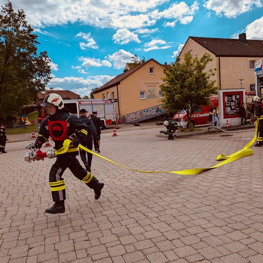 Ein Feuerwehrmann rennt mit einer Feuerwehrschläuche auf einem sonnigen Außenbereich, mit Gebäuden und Menschen im Hintergrund.